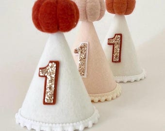 May include: Three cone-shaped party hats made of felt. Two hats are white and one is light pink. Each hat features a glittery gold number one on the front, a white pom-pom trim at the base, and a small felt pumpkin topper in orange or light pink.