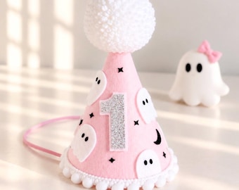 Può includere: Un cappellino da festa conico rosa chiaro con un grande numero uno in glitter argento, applicazioni di fantasmi in feltro bianco, dettagli a forma di stella e mezzaluna nera, un pon pon bianco in cima, bordo a pon pon e un sottile elastico rosa.