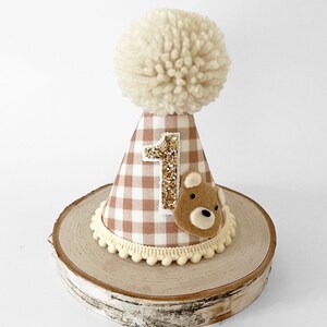 May include: A brown and white gingham fabric party hat with a white pom pom on top. The hat has a gold glitter number "1" and a brown felt bear applique. The hat is sitting on a wooden slice.