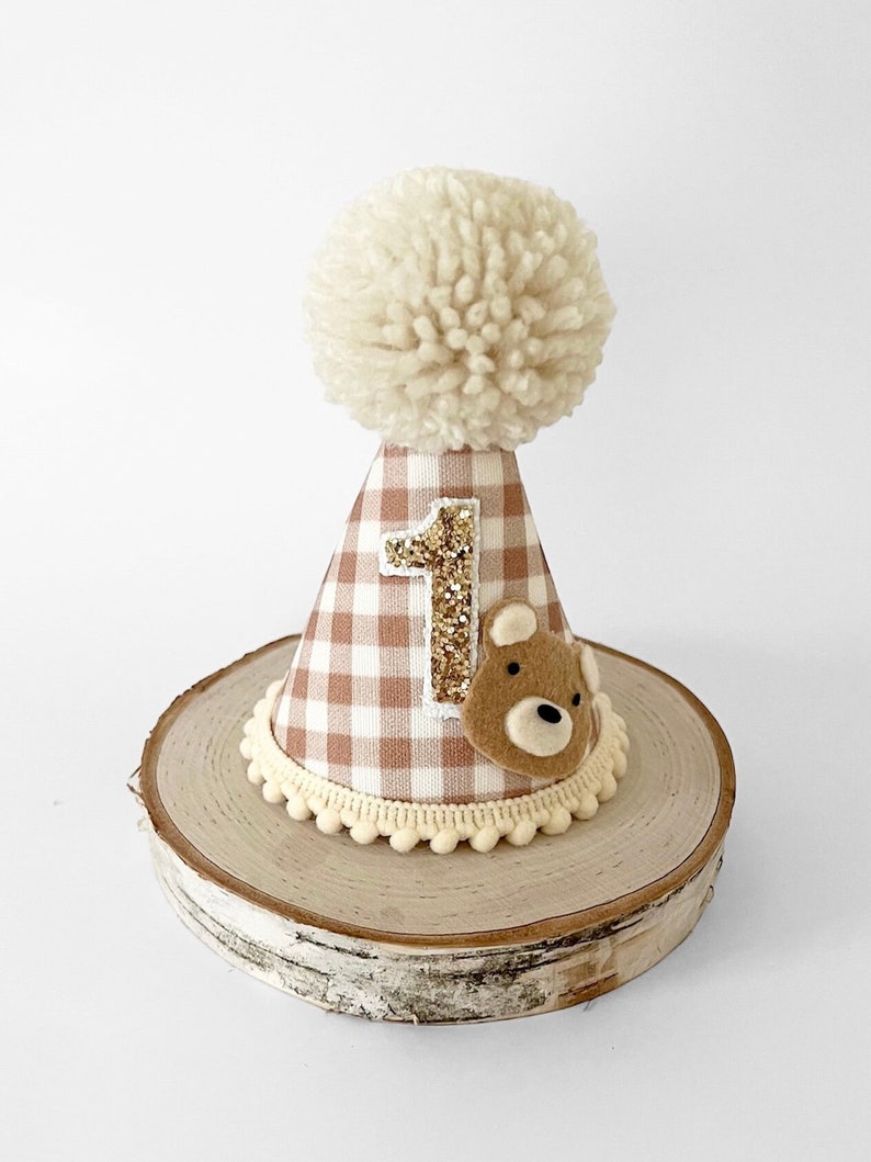 May include: A brown and white gingham fabric party hat with a white pom pom on top. The hat has a gold glitter number "1" and a brown felt bear applique. The hat is sitting on a wooden slice.