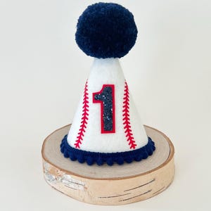 May include: White felt party hat with a blue pom-pom on top. The hat has a red baseball stitch design and a glittery black number "1" on the front.
