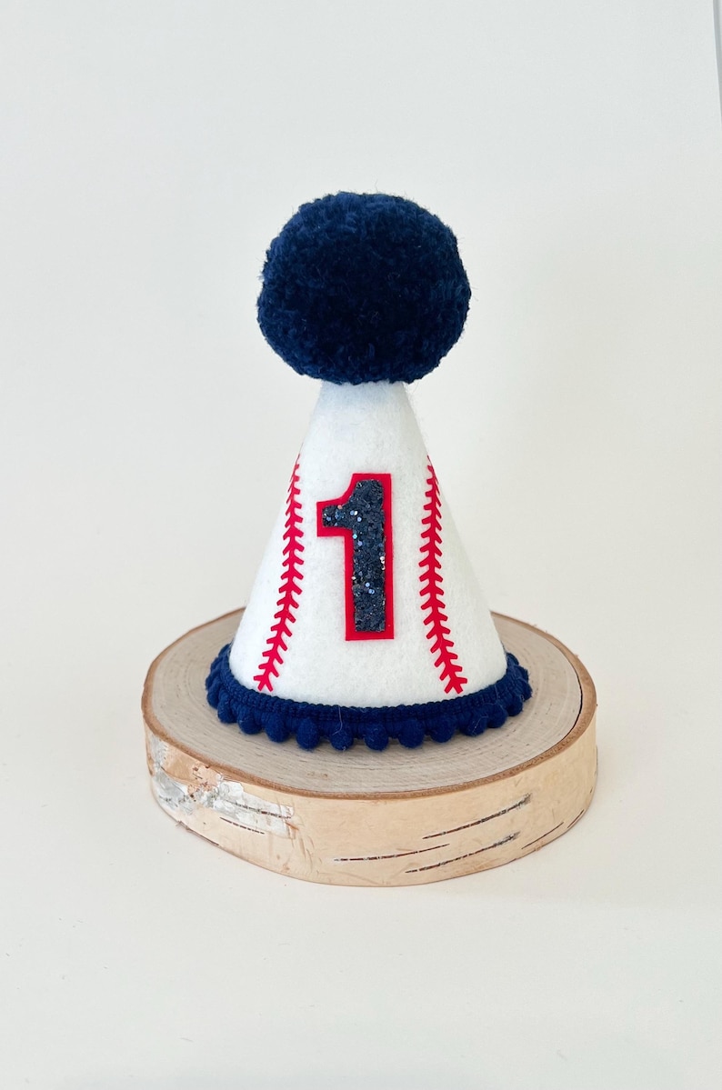 May include: White felt party hat with a blue pom-pom on top. The hat has a red baseball stitch design and a glittery black number "1" on the front.