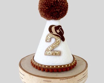 May include: White felt party hat with a brown pom-pom on top. The hat has a brown felt cowboy hat with a gold star on it and a gold glitter number "2" on the front. The hat is sitting on a wooden base.