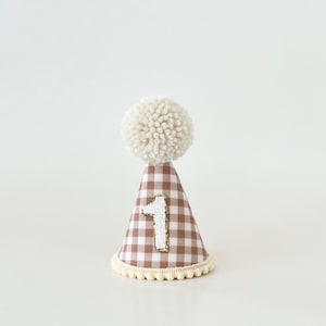 May include: A brown and white gingham party hat with a white pom-pom on top and a white felt number "1" on the front. The hat has a white trim around the bottom.