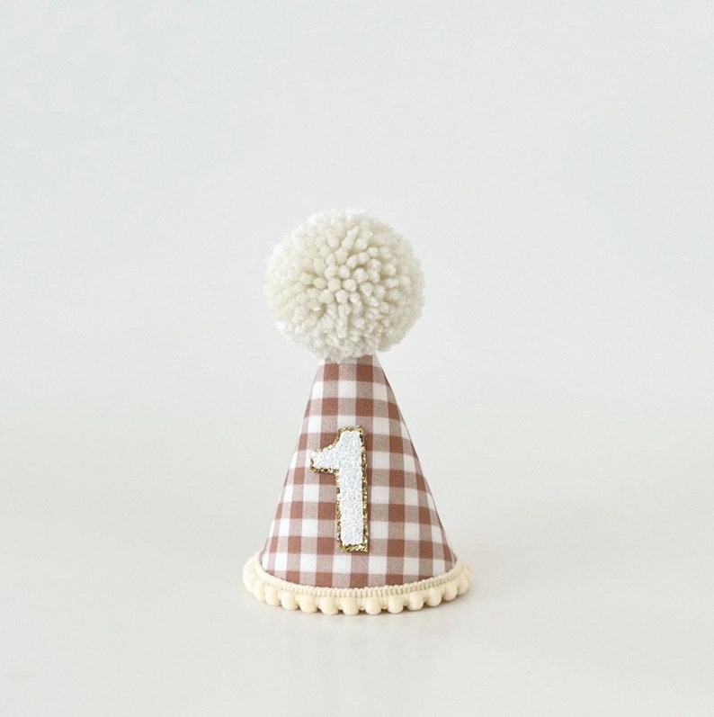 May include: A brown and white gingham party hat with a white pom-pom on top and a white felt number "1" on the front. The hat has a white trim around the bottom.