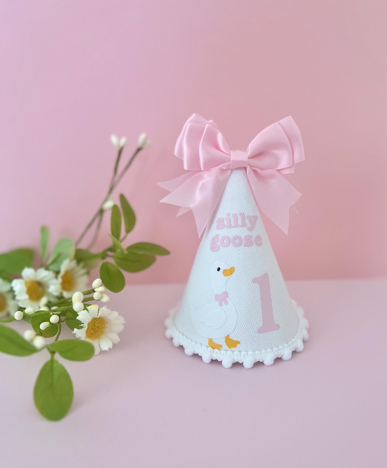 May include: A white party hat with a pink bow and trim. The hat features a cartoon goose illustration, the words "silly goose", and the number "1" in pink. The hat is decorated with a white pom-pom trim and is set against a pink background.