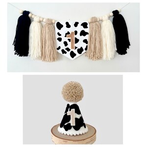 May include: A black and white cow print party banner with a gold number one. The banner is strung with white, black, and tan tassels. A matching cow print party hat with a gold number one is also shown.
