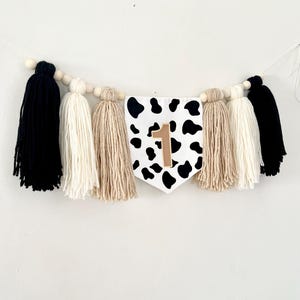 May include: A black and white cow print fabric banner with a gold number one on it. The banner is strung with wooden beads and yarn tassels in black, white, and beige.