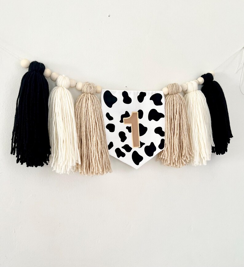 May include: A black and white cow print fabric banner with a gold number one on it. The banner is strung with wooden beads and yarn tassels in black, white, and beige.
