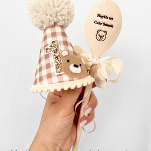 May include: A brown and white gingham first birthday hat with a white pom pom on top and a gold glitter number one. A wooden spoon with the text "Hayk's 1st Cake Smash" and a small bear illustration is included.