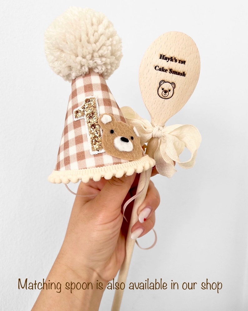 May include: A brown and white gingham first birthday hat with a white pom pom on top and a gold glitter number one. A wooden spoon with the text "Hayk's 1st Cake Smash" and a small bear illustration is included.