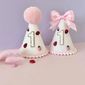 May include: Two white cone-shaped party hats decorated with small red ladybug embellishments and a glittery gold number one. One hat features a large pink pom-pom on top, while the other is adorned with a light pink satin ribbon bow.