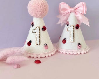May include: Two white cone-shaped party hats decorated with small red ladybug embellishments and a glittery gold number one. One hat features a large pink pom-pom on top, while the other is adorned with a light pink satin ribbon bow.