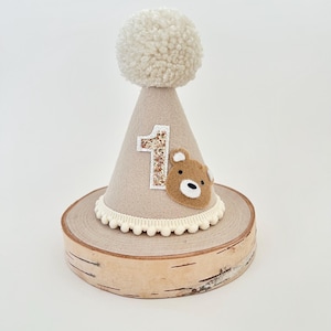 May include: A beige felt cone-shaped party hat with a white pom-pom on top. The hat has a brown felt bear applique and a gold glitter number "1" on the front. The hat is sitting on a wooden base.