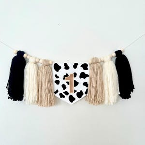 May include: A black and white cow print banner with a gold number one on it. The banner is strung with wooden beads and has black, white, and beige tassels.