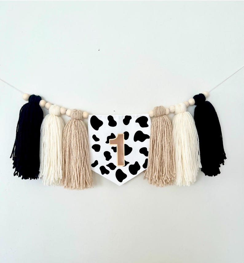 May include: A black and white cow print banner with a gold number one on it. The banner is strung with wooden beads and has black, white, and beige tassels.