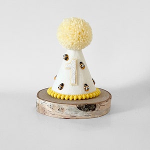 May include: White felt party hat with a yellow pom-pom on top. The hat is decorated with yellow pom-poms around the base and black and yellow felt bees. The number "1" is in the center of the hat.