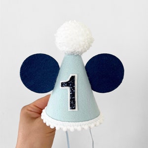 May include: A light blue felt party hat with a white pom-pom on top and blue felt ears. The hat has a white trim and a white string attached. The number "1" is on the front of the hat in black glitter.