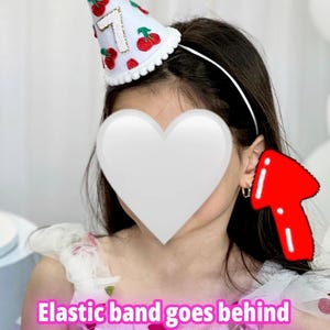 May include: A white party hat with a fluffy pom-pom and a scalloped edge. The hat is decorated with red cherries and the number 7 in gold. The hat is attached to a white headband.