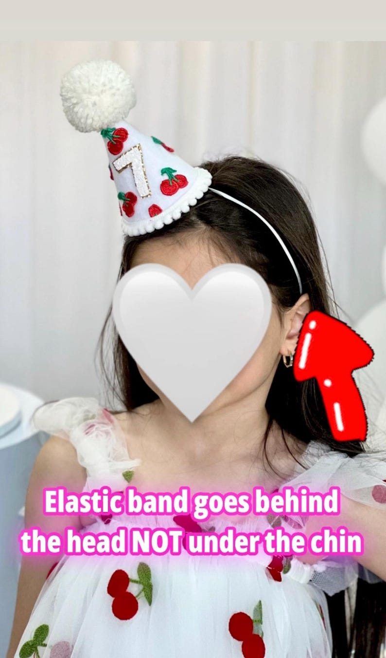 May include: A white party hat with a fluffy pom-pom and a scalloped edge. The hat is decorated with red cherries and the number 7 in gold. The hat is attached to a white headband.