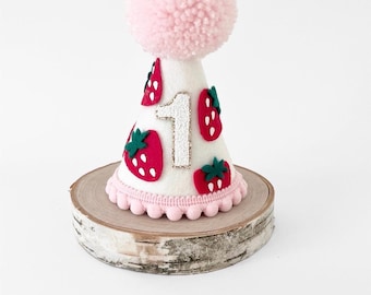 May include: A white cone-shaped party hat featuring red strawberry appliqués with green leaves and a glittery silver number one on the front. The hat is topped with a large light pink yarn pom-pom and finished with a light pink pom-pom trim.