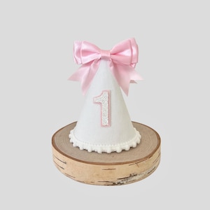May include: White fabric party hat with a pink bow and the number "1" on the front. The hat is sitting on a wooden base.