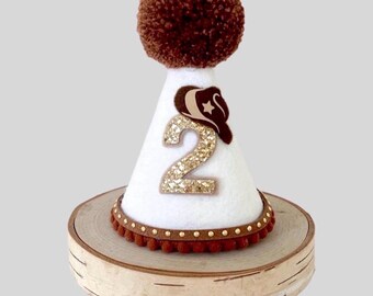 May include: A white cone-shaped party hat featuring a large brown pom-pom on top, a gold glittery number two, and a brown cowboy hat applique. The base is trimmed with a brown band featuring small gold studs and a brown pom-pom fringe.