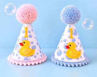 May include: Two cone-shaped party hats featuring a light blue bubble pattern on a white background. Each hat displays a yellow rubber duck applique, a yellow number one, and a pom-pom topper. One hat has pink accents, while the other has blue.