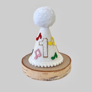 May include: White felt party hat with a white pom-pom on top. The hat has a number "1" on the front with glitter and colorful musical notes around it. The hat is sitting on a wooden base.