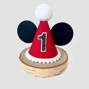 May include: A red cone-shaped party hat featuring a white pom-pom on top, a white pom-pom trim at the base, and two black circular ears on the sides. The front of the hat displays a black glittery number one with a white border.