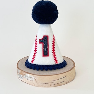 May include: White felt cone party hat with a red and white baseball design. The hat has a blue pom-pom on top and a blue trim with white pom-poms. The number "1" is on the front of the hat in black glitter.