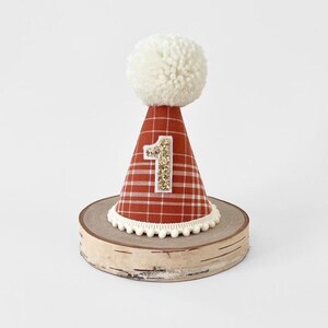 May include: A red and white plaid party hat with a white pom pom on top. The hat has a gold glitter number "1" on the front. The hat is sitting on a wooden base.