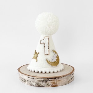 May include: White felt party hat with a gold glitter number one, a gold glitter moon with white stars, and a gold glitter star. The hat has a white pom-pom on top and a white scalloped edge.