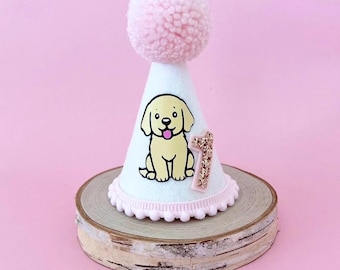 May include: White felt party hat with a pink pom pom on top. The hat has a yellow puppy illustration and a gold glitter number one on the front. The hat is sitting on a wooden base.