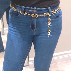 May include: A gold-colored chain belt with a decorative design. The belt is worn over blue jeans and features a series of connected links and a dangling charm. The belt adds a touch of elegance to the casual outfit.