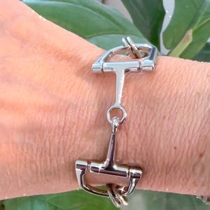 May include: Silver-toned equestrian-style bracelet featuring a snaffle bit design. The bracelet is made of polished metal and has a classic, equestrian theme. The design includes a central bit and connecting links.