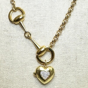 May include: A gold-toned necklace featuring a horse bit design and a heart-shaped pendant. The heart pendant is encrusted with small, clear crystals. The necklace has a chain with a round link design.