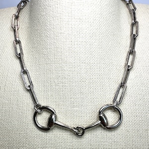 May include: Silver-toned necklace featuring a chain of rectangular links and a horse bit design. The necklace is displayed on a neutral-colored surface, highlighting the metallic sheen of the jewelry. The horse bit is a prominent focal point.