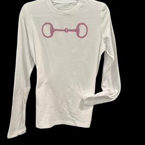 May include: White long-sleeve t-shirt with a pink glitter horse bit design. The shirt has the text "Equestrians" on the neckline.