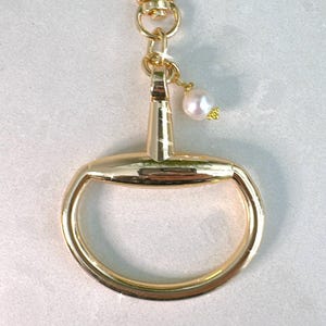 May include: Gold-colored keychain with a large, rounded ring and a smaller, rectangular top. A small, white pearl charm hangs from the top. The keychain has a lobster clasp for easy attachment.