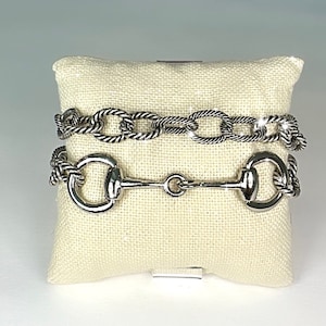 May include: Two silver bracelets on a white fabric display stand. One bracelet is a thick chain with a rope-like texture. The other bracelet is a thinner chain with a horse bit design.