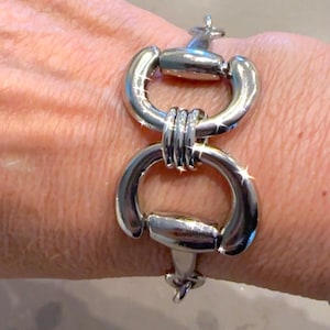May include: A silver-toned bracelet with a unique design. The bracelet features two large, connected, curved links, each with a decorative element. The links are joined by smaller, textured metal pieces. The bracelet is worn on a wrist.
