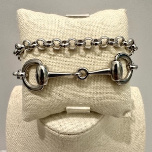 May include: Two silver bracelets displayed on a beige linen surface. The top bracelet has a chain link design. The bottom bracelet features a horse bit design with two circular rings and a central bar.