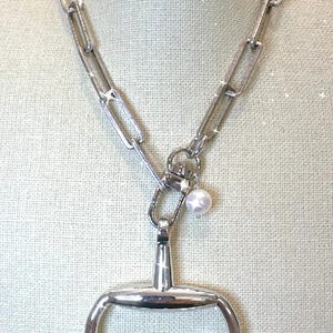 May include: Silver-toned necklace featuring a large link chain and a horse bit pendant. A small pearl charm hangs from the clasp. The necklace is displayed on a neutral-toned background.