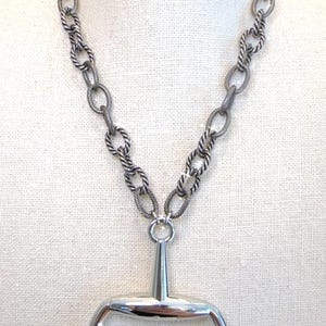 May include: Silver-toned necklace featuring a horse bit pendant. The necklace has a textured, twisted chain design with a large oval pendant. The pendant is a polished silver color, and the necklace is displayed on a white surface.