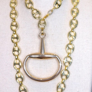 May include: A gold-toned necklace featuring a horse bit pendant and a chain with oval links. The necklace is displayed on a white surface, showcasing its shiny, reflective surface. The jewelry is a statement piece.