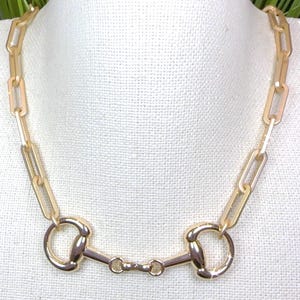 May include: Gold-toned necklace featuring a horse bit design. The necklace has a chain of rectangular links and a central horse bit element with two rounded rings connected by a bar. The jewelry is displayed on a white surface.