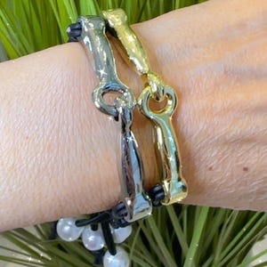 May include: Two metal bracelets, one silver and one gold, are worn on a wrist. The bracelets have a unique hook-and-eye design and are attached to a black leather band. White pearl beads are also attached to the band.