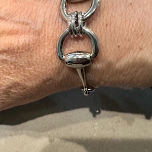 May include: Silver-toned bracelet featuring a series of linked, rounded shapes. The bracelet has a unique design with a bird-like element at the top of the clasp. The bracelet is a stylish accessory.