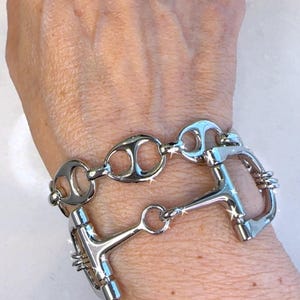 May include: A silver-toned bracelet featuring a horse bit design. The bracelet is composed of multiple connected links, including oval and rectangular shapes, and is worn on a wrist. The metal reflects light, creating a shiny appearance.